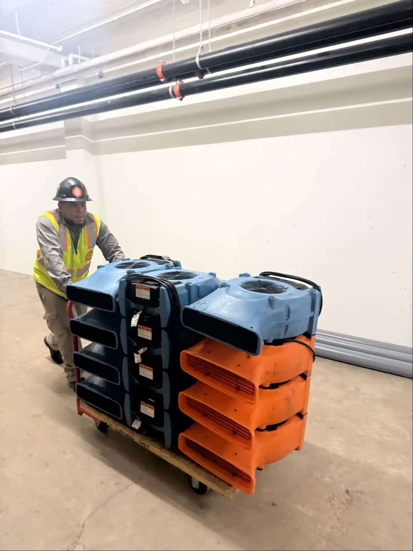 Worker in a hard hat pushing a cart stacked with air movers across a garage